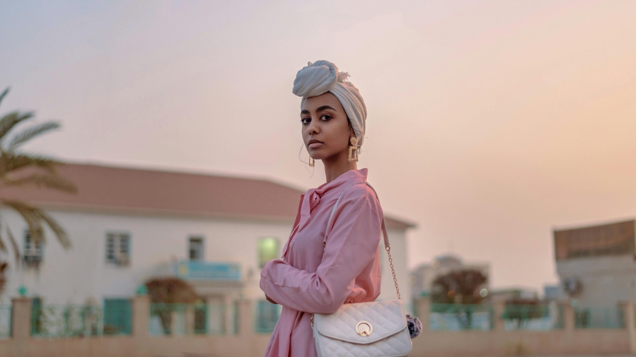 Stylish woman in pink dress with turban, posing outdoors at sunset.
