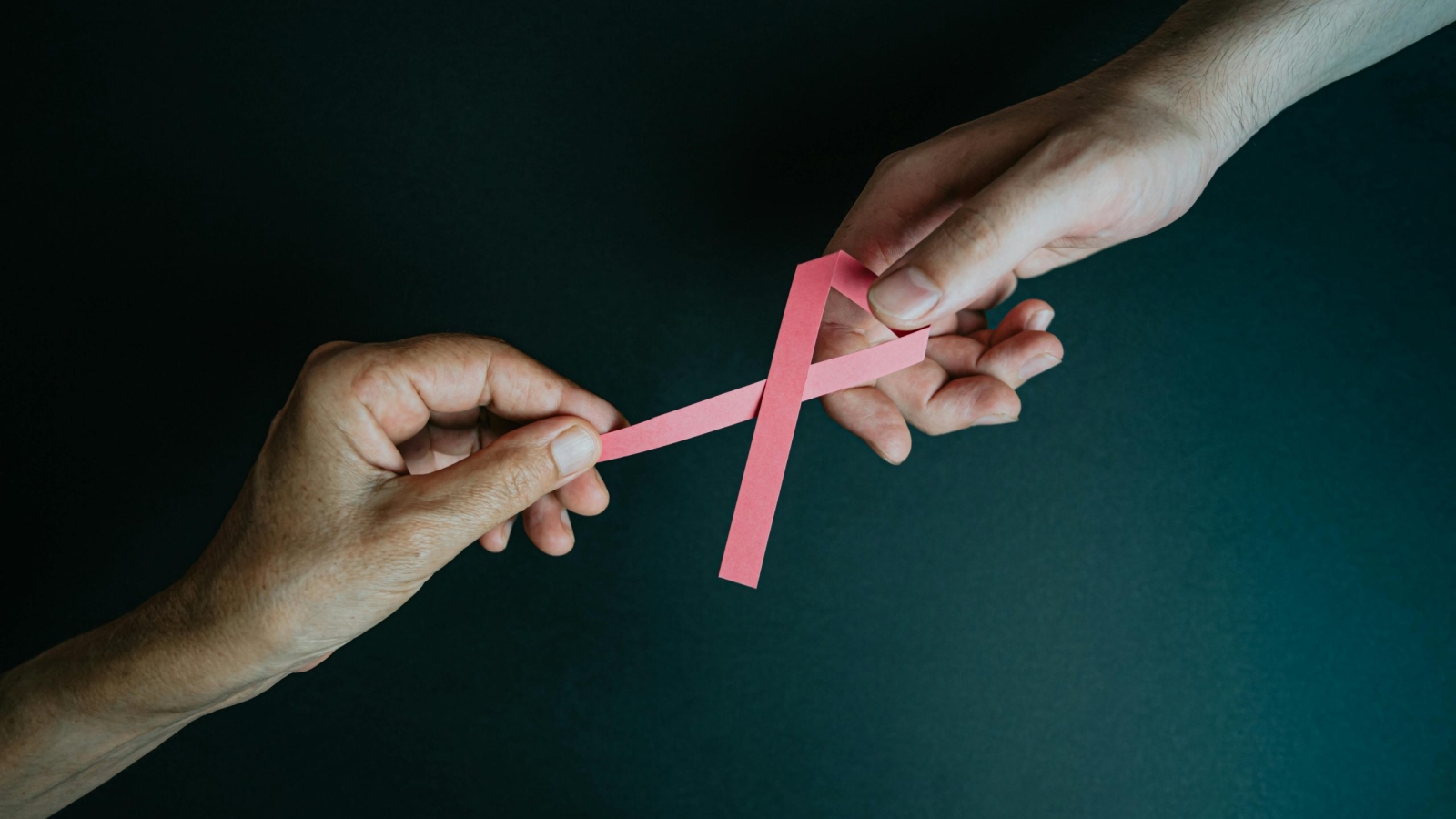 Two hands exchanging a pink ribbon symbolizing breast cancer awareness and support.